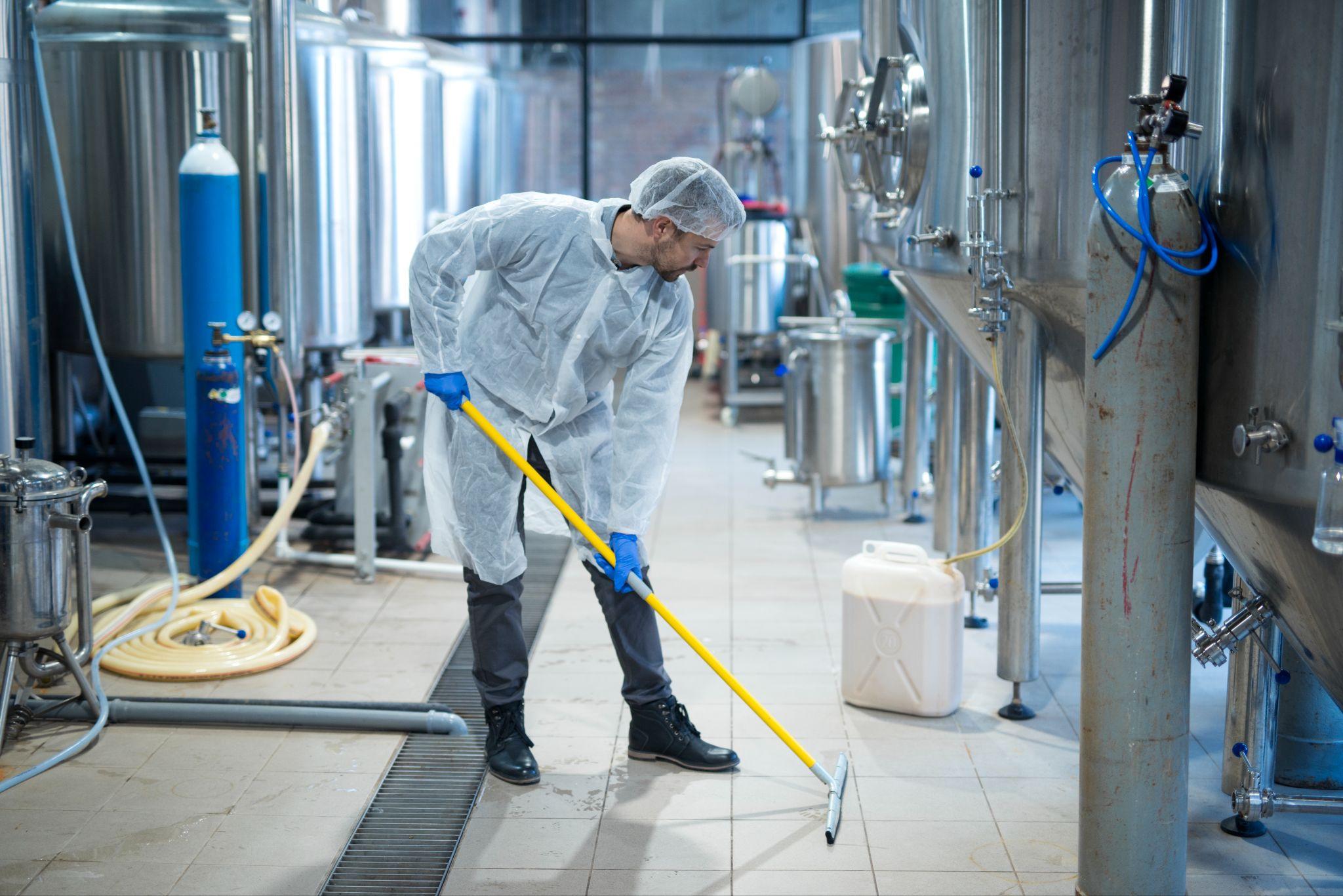 A man wearing a cleaning uniform mops the floor of an industrial room. The room appears to have specialized equipment and machinery